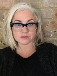 Image of a caucasian woman with blonde hair in a bob and spectacles staring straight at the camera
